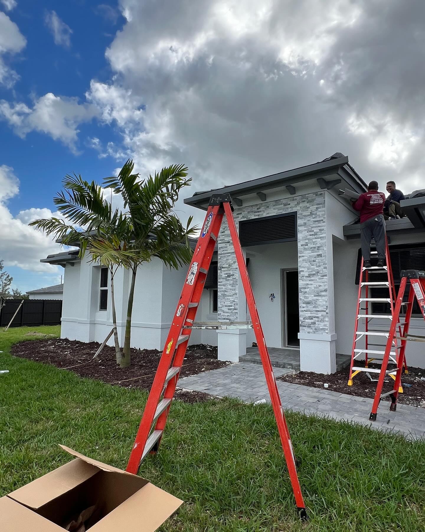 Crew on ladders installs gutters on a new stone-accent home; tools in yard.