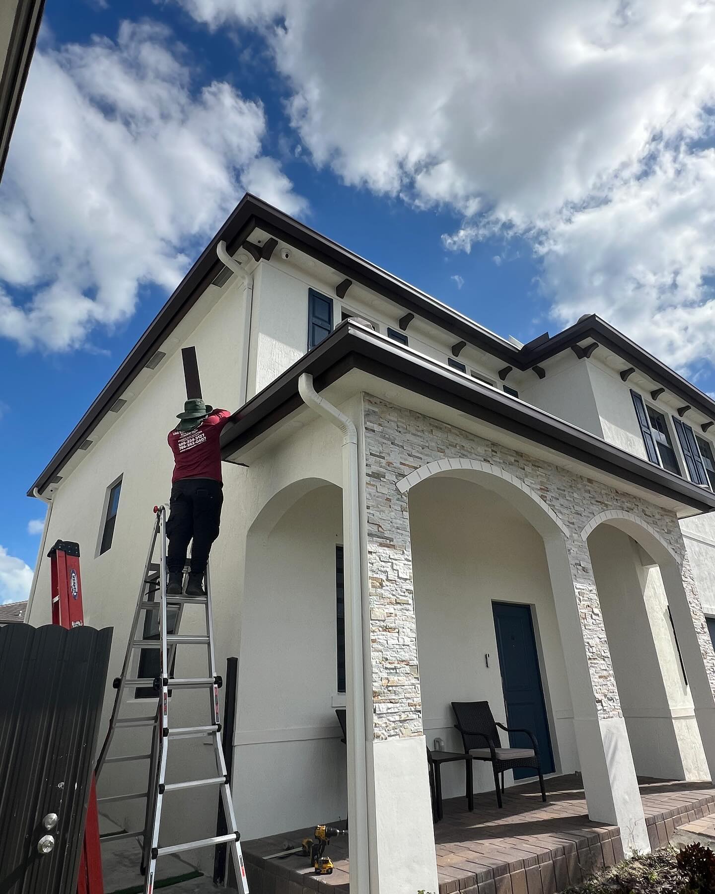 Worker on a ladder installs brown gutter on a two-story stone-and-stucco home.