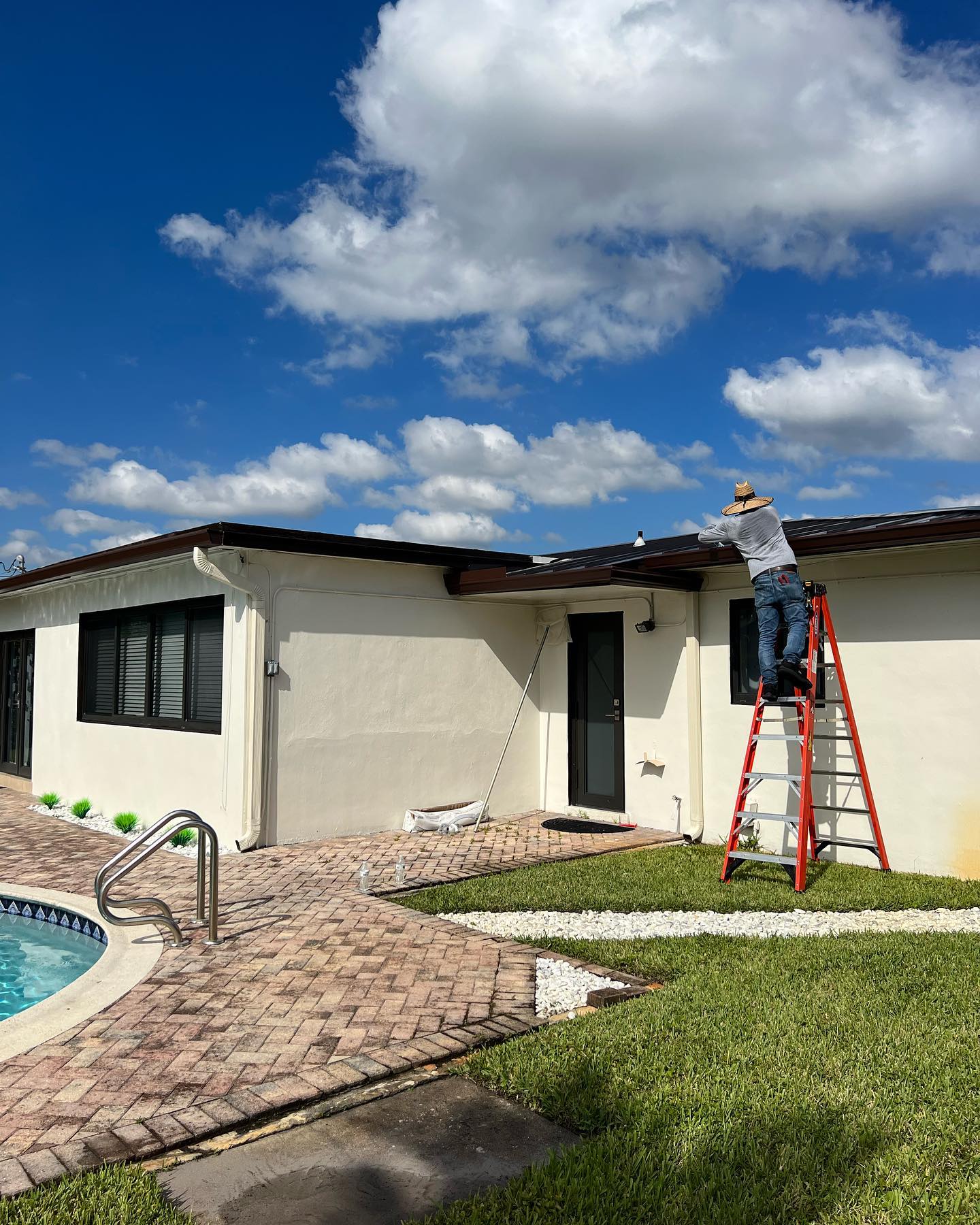 Worker on a ladder installing gutter along a single-story home beside a pool.