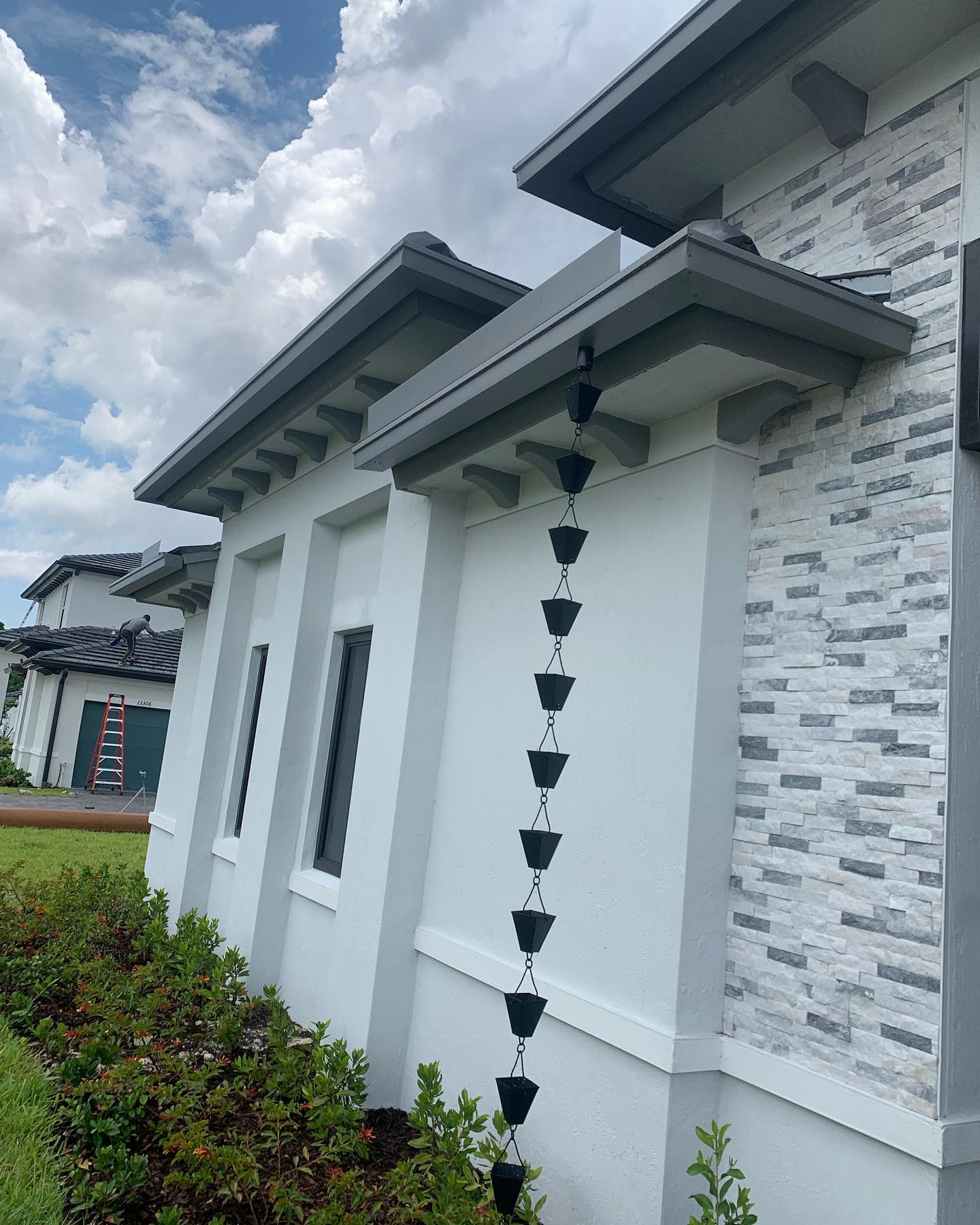 Black cup rain chain on white stone-and-stucco home with crew on ladder in background.
